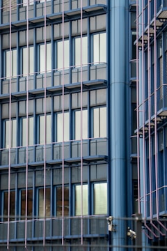 A modern building facade featuring a grid of reflective windows framed with blue, vertical and horizontal panels. The structure includes glass balconies and security cameras at various points. The reflections in the windows suggest an urban environment.