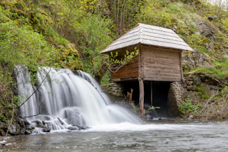 A small wooden structure sits beside a gentle waterfall, surrounded by lush greenery and moss-covered rocks. The rustic wooden building is elevated on stone supports, blending harmoniously with the natural landscape.