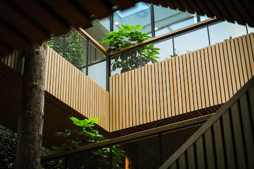 An interior space featuring wooden architectural elements with slatted walls and railings. Green foliage adds a natural touch, with large plants appearing on both levels. Natural light filters through a skylight above, creating a serene atmosphere.