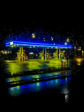 A gas station is brightly illuminated at night with neon lights reflecting off a rain-soaked surface. The droplets on a window create a textured effect, with blue and yellow lights casting a vibrant glow.