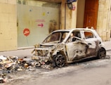 A burnt-out car is parked next to a pile of trash on an urban street. The car's exterior is charred and stripped, with no windows or front bumper remaining. Debris and ashes are scattered around the area, indicating a fire occurred. A glass window behind the car has a colorful directional sign with French text and some graffiti.