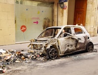 A burnt-out car is parked next to a pile of trash on an urban street. The car's exterior is charred and stripped, with no windows or front bumper remaining. Debris and ashes are scattered around the area, indicating a fire occurred. A glass window behind the car has a colorful directional sign with French text and some graffiti.