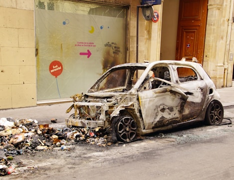 A burnt-out car is parked next to a pile of trash on an urban street. The car's exterior is charred and stripped, with no windows or front bumper remaining. Debris and ashes are scattered around the area, indicating a fire occurred. A glass window behind the car has a colorful directional sign with French text and some graffiti.