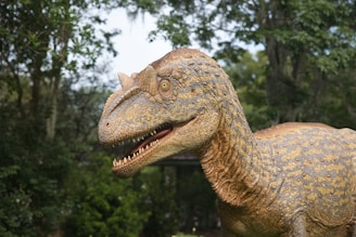 A detailed, close-up view of a dinosaur model set against a backdrop of lush, green trees. The dinosaur features textured, scaly skin with a blend of orange, brown, and gray colors. Its mouth is open, displaying sharp, white teeth, and its eyes are round and striking.