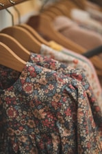 Several wooden hangers hold a variety of floral-patterned clothing items, with the focus on a richly patterned dress or blouse in the foreground. The hanging garments display a mixture of colorful flowers in shades of red, orange, and blue on darker backgrounds.