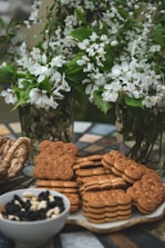 A collection of intricately designed cookies and oat biscuits are positioned on a decorative table. Behind them, lush green branches adorned with delicate white flowers are arranged in glass vases, creating a fresh and lively backdrop. Nearby, a bowl filled with a mix of dried fruits and nuts adds to the assortment. The setting evokes a warm, inviting atmosphere.