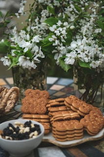 A collection of intricately designed cookies and oat biscuits are positioned on a decorative table. Behind them, lush green branches adorned with delicate white flowers are arranged in glass vases, creating a fresh and lively backdrop. Nearby, a bowl filled with a mix of dried fruits and nuts adds to the assortment. The setting evokes a warm, inviting atmosphere.