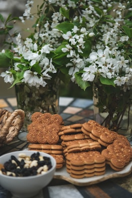 A collection of intricately designed cookies and oat biscuits are positioned on a decorative table. Behind them, lush green branches adorned with delicate white flowers are arranged in glass vases, creating a fresh and lively backdrop. Nearby, a bowl filled with a mix of dried fruits and nuts adds to the assortment. The setting evokes a warm, inviting atmosphere.