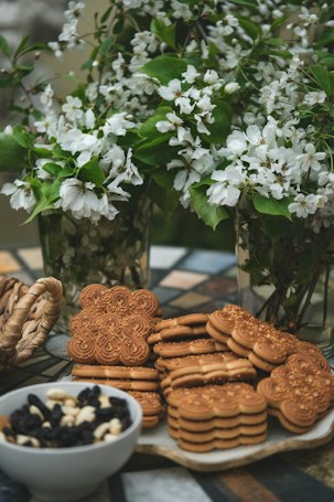A collection of intricately designed cookies and oat biscuits are positioned on a decorative table. Behind them, lush green branches adorned with delicate white flowers are arranged in glass vases, creating a fresh and lively backdrop. Nearby, a bowl filled with a mix of dried fruits and nuts adds to the assortment. The setting evokes a warm, inviting atmosphere.