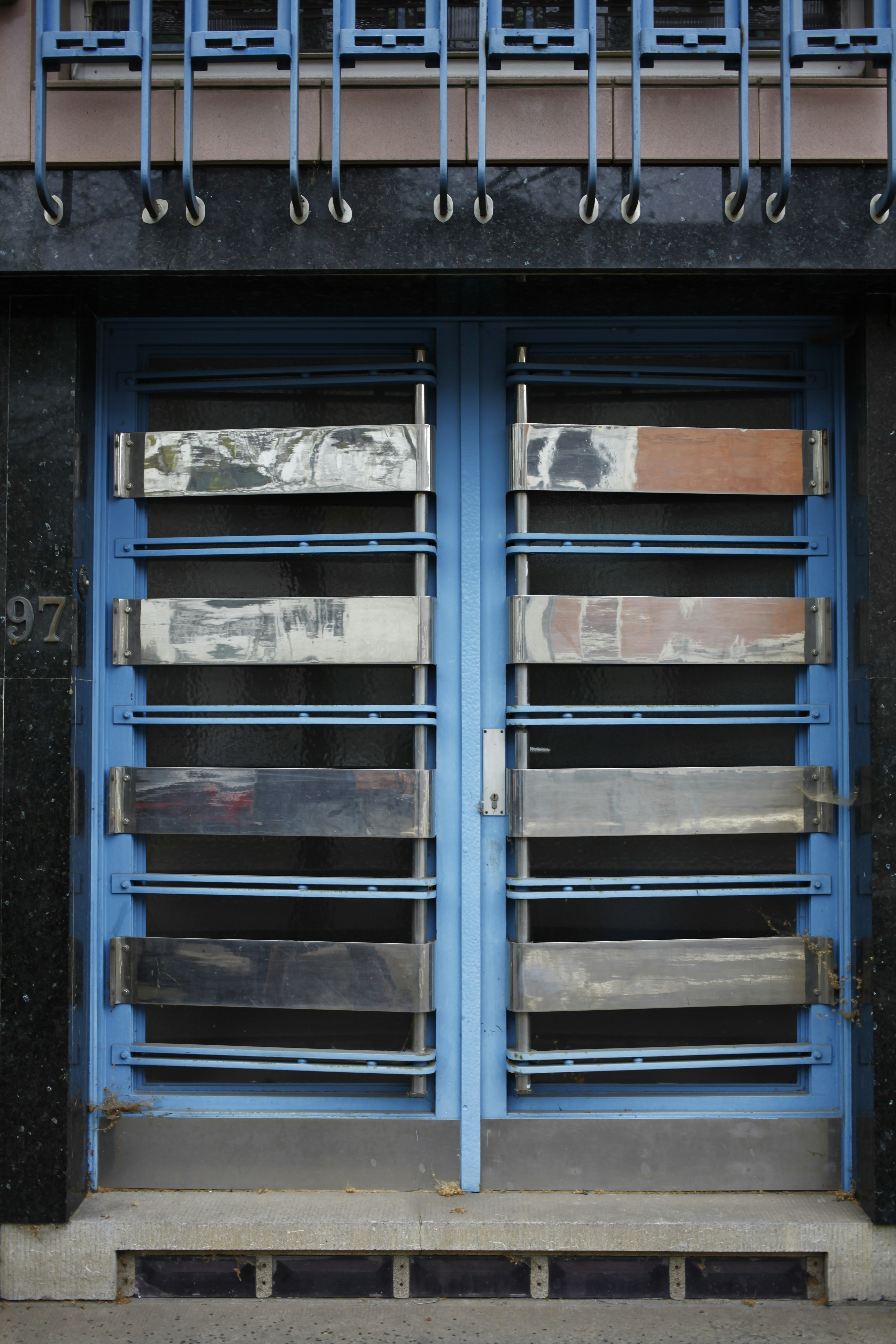 A double door painted in blue with metallic horizontal bars across each section. Above the doors, a row of vertical metal bars is attached to an overhanging section. The doors and the structure surrounding them exhibit a combination of industrial and modern design.