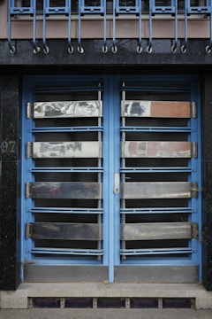 A double door painted in blue with metallic horizontal bars across each section. Above the doors, a row of vertical metal bars is attached to an overhanging section. The doors and the structure surrounding them exhibit a combination of industrial and modern design.