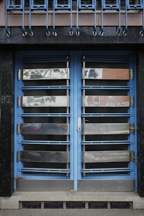 A double door painted in blue with metallic horizontal bars across each section. Above the doors, a row of vertical metal bars is attached to an overhanging section. The doors and the structure surrounding them exhibit a combination of industrial and modern design.