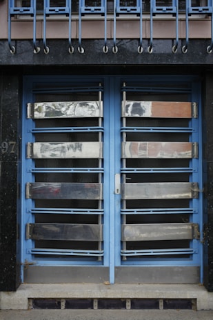 A double door painted in blue with metallic horizontal bars across each section. Above the doors, a row of vertical metal bars is attached to an overhanging section. The doors and the structure surrounding them exhibit a combination of industrial and modern design.