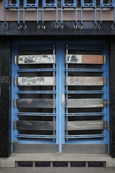 A double door painted in blue with metallic horizontal bars across each section. Above the doors, a row of vertical metal bars is attached to an overhanging section. The doors and the structure surrounding them exhibit a combination of industrial and modern design.