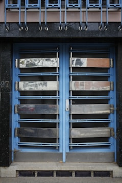 A double door painted in blue with metallic horizontal bars across each section. Above the doors, a row of vertical metal bars is attached to an overhanging section. The doors and the structure surrounding them exhibit a combination of industrial and modern design.