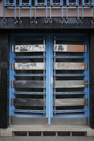 A double door painted in blue with metallic horizontal bars across each section. Above the doors, a row of vertical metal bars is attached to an overhanging section. The doors and the structure surrounding them exhibit a combination of industrial and modern design.