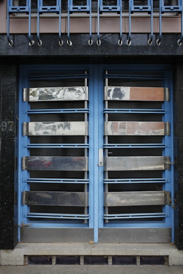 A double door painted in blue with metallic horizontal bars across each section. Above the doors, a row of vertical metal bars is attached to an overhanging section. The doors and the structure surrounding them exhibit a combination of industrial and modern design.