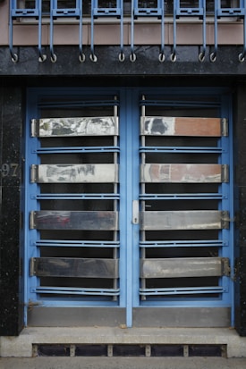 A double door painted in blue with metallic horizontal bars across each section. Above the doors, a row of vertical metal bars is attached to an overhanging section. The doors and the structure surrounding them exhibit a combination of industrial and modern design.