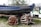 A blue boat named 'Tender te Potvis' is placed on a trailer with a cobblestone ground in the foreground. Next to the boat, several orange and white lobster pots are stacked and tied with ropes. The background shows green grass and some bushes, along with partial views of other watercraft.
