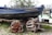 A blue boat named 'Tender te Potvis' is placed on a trailer with a cobblestone ground in the foreground. Next to the boat, several orange and white lobster pots are stacked and tied with ropes. The background shows green grass and some bushes, along with partial views of other watercraft.