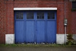 A pair of large blue wooden doors set into a red brick wall, featuring rectangular windows at the top. The surrounding brickwork shows some signs of weathering, and there is a small patch of green moss growing at the base of the wall. An electrical meter is mounted on the right side near the door.