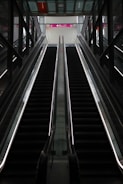 An empty escalator in a modern indoor setting ascends towards a brightly lit area with glass and metal surroundings. Overhead signs indicate directions for various locations.