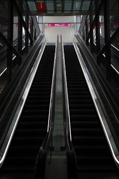An empty escalator in a modern indoor setting ascends towards a brightly lit area with glass and metal surroundings. Overhead signs indicate directions for various locations.