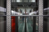 A symmetrical view through a glass window framed with metallic panels featuring a diamond plate pattern. The background appears to be a modern corridor or hallway, with blurred elements suggesting depth and perspective.