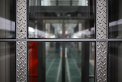 A symmetrical view through a glass window framed with metallic panels featuring a diamond plate pattern. The background appears to be a modern corridor or hallway, with blurred elements suggesting depth and perspective.