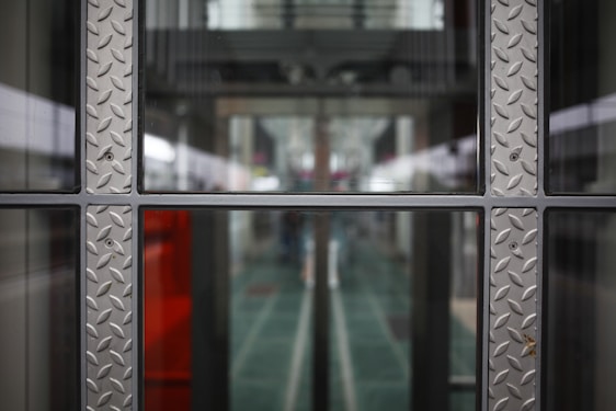 A symmetrical view through a glass window framed with metallic panels featuring a diamond plate pattern. The background appears to be a modern corridor or hallway, with blurred elements suggesting depth and perspective.
