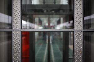 A symmetrical view through a glass window framed with metallic panels featuring a diamond plate pattern. The background appears to be a modern corridor or hallway, with blurred elements suggesting depth and perspective.