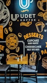 A coffee shop interior features a decorative wall with typography and graphics related to coffee and desserts. The wall displays phrases and images of coffee cups, coffee beans, and desserts like cupcakes and cheesecake. In front of the wall, a wooden table with two cushioned chairs is set up, accompanied by a small ashtray and a metal cup.