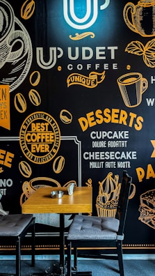 A coffee shop interior features a decorative wall with typography and graphics related to coffee and desserts. The wall displays phrases and images of coffee cups, coffee beans, and desserts like cupcakes and cheesecake. In front of the wall, a wooden table with two cushioned chairs is set up, accompanied by a small ashtray and a metal cup.