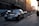 A luxury black sedan is parked on the side of a cobblestone street in an urban area. The sun is setting, casting a warm glow on the buildings, which feature historic architecture with intricate details. Several other vehicles are visible in the background as they drive along the street.