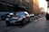 A luxury black sedan is parked on the side of a cobblestone street in an urban area. The sun is setting, casting a warm glow on the buildings, which feature historic architecture with intricate details. Several other vehicles are visible in the background as they drive along the street.