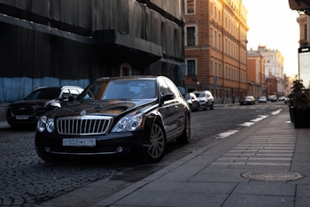 A luxury black sedan is parked on the side of a cobblestone street in an urban area. The sun is setting, casting a warm glow on the buildings, which feature historic architecture with intricate details. Several other vehicles are visible in the background as they drive along the street.