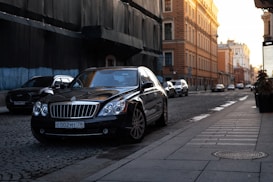 A luxury black sedan is parked on the side of a cobblestone street in an urban area. The sun is setting, casting a warm glow on the buildings, which feature historic architecture with intricate details. Several other vehicles are visible in the background as they drive along the street.