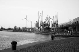 A black and white image of an industrial waterfront featuring several large cranes along the dock. In the background, wind turbines are visible. The cobblestone pavement leads the viewer's eye along the waterfront. Buildings with a modern design can be seen on the right side.