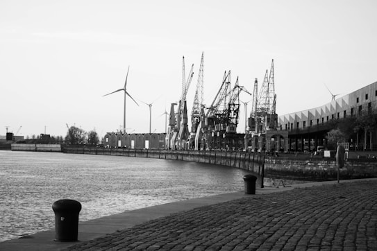 A black and white image of an industrial waterfront featuring several large cranes along the dock. In the background, wind turbines are visible. The cobblestone pavement leads the viewer's eye along the waterfront. Buildings with a modern design can be seen on the right side.