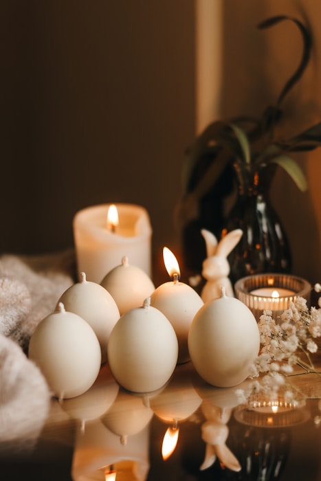 A cozy arrangement of round candles partially lit, accompanied by a small figurine resembling a rabbit and a delicate sprig of dried flowers, set upon a reflective surface. In the background, an additional lit candle and a dark vase with green leaves add to the serene ambiance.