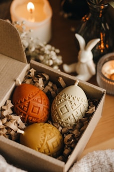 Three intricately designed candles shaped like Easter eggs are placed in a small cardboard box filled with shredded paper. The candles are colored orange, cream, and yellow, and have geometric patterns etched into them. In the background, a lit candle, a white ceramic bunny, and some decorative flowers are visible, creating a cozy and warm atmosphere.
