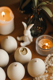 A cozy arrangement featuring a collection of white round candles, a ceramic rabbit figure, and a dark glass vase with leafy foliage on a wooden surface. Two lit candles, one in a glass holder and one in a ceramic cup, add a warm and intimate glow. Delicate white flowers complement the scene.
