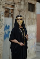 A person wearing a traditional black outfit adorned with a gold coin face veil, standing against a weathered stone wall with graffiti. The attire includes a black robe with gold necklaces and bracelets, creating an elegant and mysterious appearance.