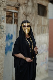 A person wearing a traditional black outfit adorned with a gold coin face veil, standing against a weathered stone wall with graffiti. The attire includes a black robe with gold necklaces and bracelets, creating an elegant and mysterious appearance.