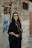 A person wearing a traditional black outfit adorned with a gold coin face veil, standing against a weathered stone wall with graffiti. The attire includes a black robe with gold necklaces and bracelets, creating an elegant and mysterious appearance.
