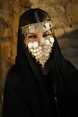 A person is adorned in a black garment and wearing an intricate gold face mask with decorative coins covering the lower half of the face. The eyes are prominently visible through the mask. The background features a textured stone wall, creating a contrast with the dark clothing and the golden embellishments.