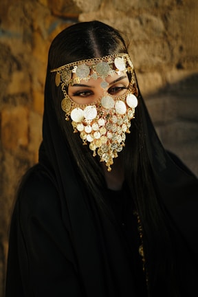 A person is adorned in a black garment and wearing an intricate gold face mask with decorative coins covering the lower half of the face. The eyes are prominently visible through the mask. The background features a textured stone wall, creating a contrast with the dark clothing and the golden embellishments.