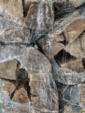 A collection of chopped firewood stacked together, partially covered with a thin, transparent, and worn plastic sheet. The pieces of wood vary in size and texture, displaying rugged edges and a natural brown hue. The plastic sheet is crinkled and covered with a dusty residue, creating an overlay effect on the wood textures.