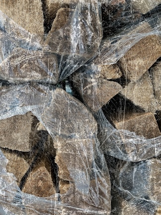 A collection of chopped firewood stacked together, partially covered with a thin, transparent, and worn plastic sheet. The pieces of wood vary in size and texture, displaying rugged edges and a natural brown hue. The plastic sheet is crinkled and covered with a dusty residue, creating an overlay effect on the wood textures.