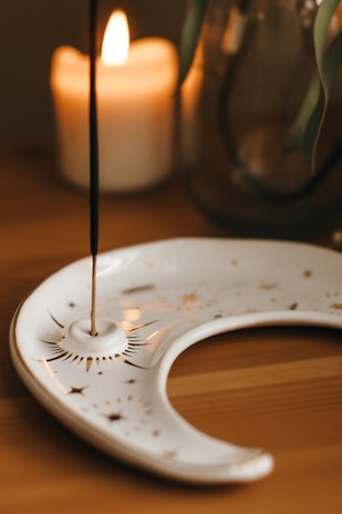 A lit candle is placed on a wooden surface next to a decorative incense holder shaped like a crescent moon. The incense stick is vertical, emitting a thin trail of smoke. In the background, there is a glass jar with plant leaves.
